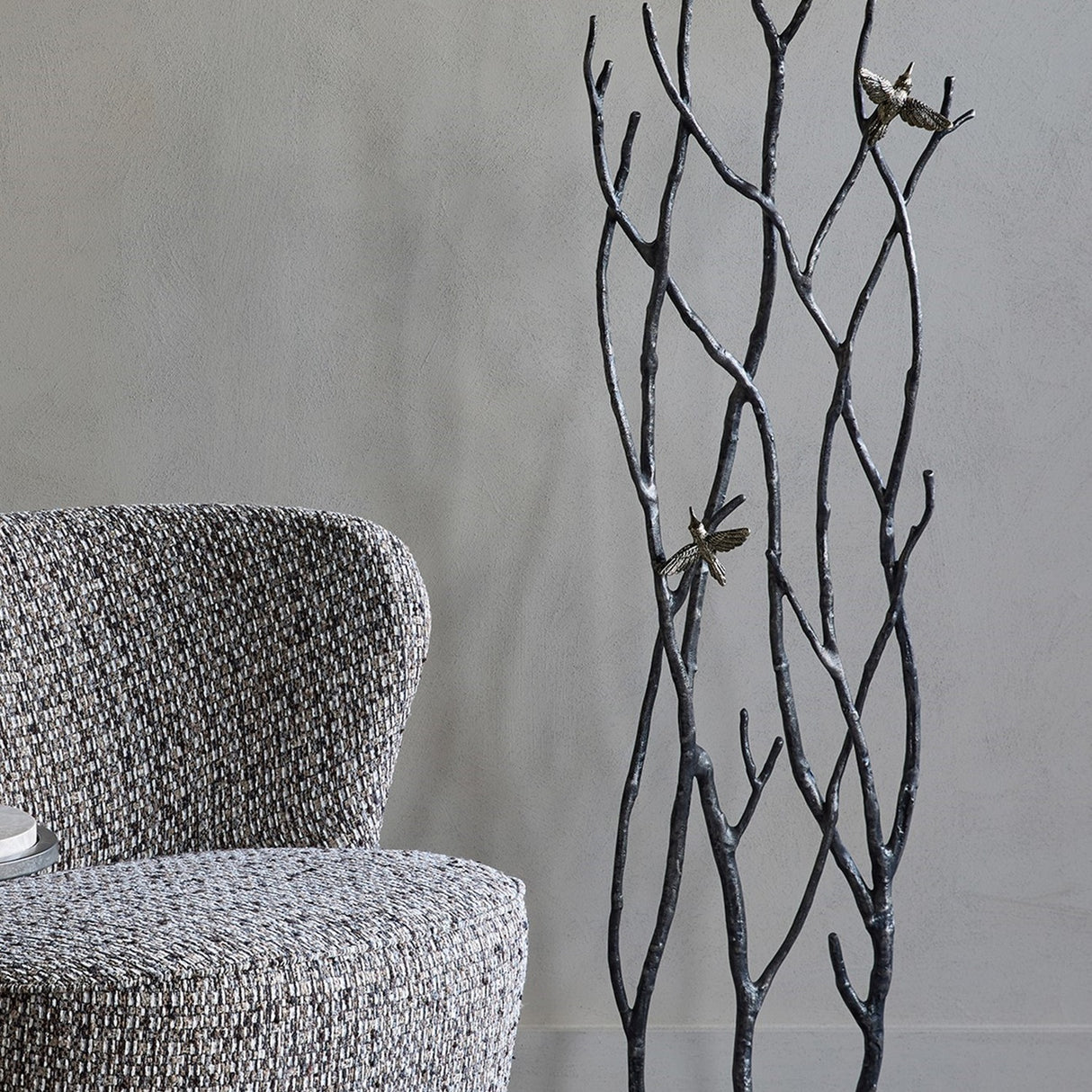 Libra Interiors Aluminium Branch and Bird Floor Sculpture in matt black with two antique brass birds, styled beside a grey textured armchair.
