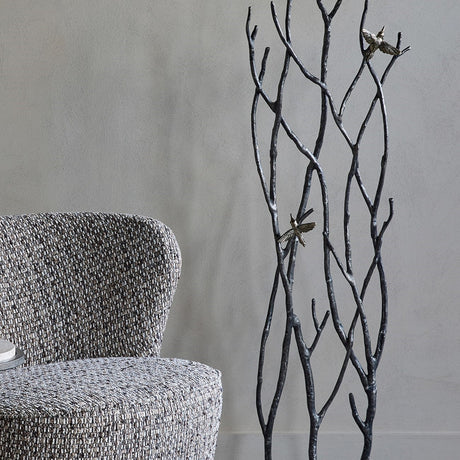 Libra Interiors Aluminium Branch and Bird Floor Sculpture in matt black with two antique brass birds, styled beside a grey textured armchair.