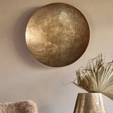 Libra Interiors Round Textured Aluminium Wall Art in antique brass, hung on a neutral wall above a brass vase with dried palm leaves.