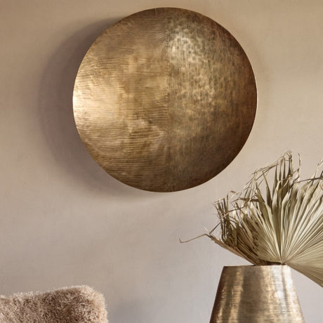 Libra Interiors Round Textured Aluminium Wall Art in antique brass, hung on a neutral wall above a brass vase with dried palm leaves.