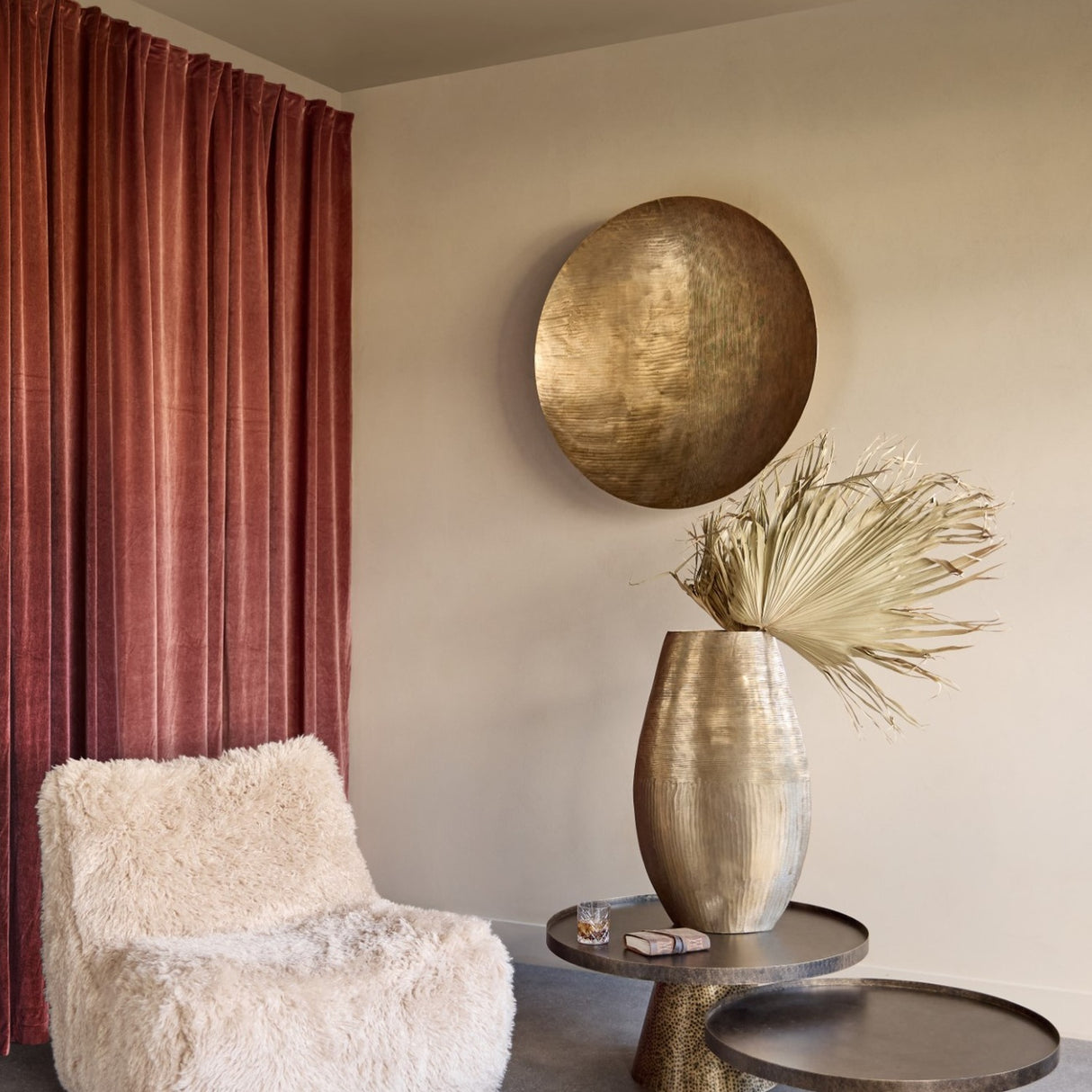 Libra Interiors Round Textured Aluminium Wall Art in antique brass above a hammered vase and side tables by a rust velvet curtain and sheepskin chair.
