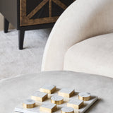 Libra Interiors Marble Effect White and Brass Noughts and Crosses Game on a grey ottoman, with a cream curved armchair and dark cabinet behind.
