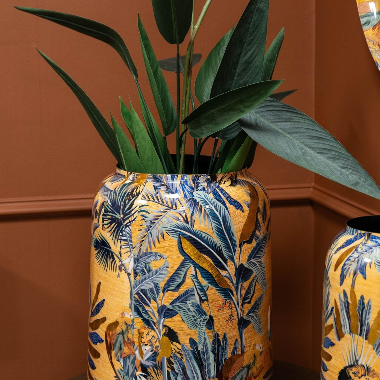 Libra Interiors gold Painted Iron Jungle Planter with tropical foliage and animal motifs, holding a tall leafy plant against a terracotta wall.