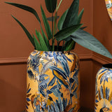 Libra Interiors gold Painted Iron Jungle Planter with tropical foliage and animal motifs, holding a tall leafy plant against a terracotta wall.
