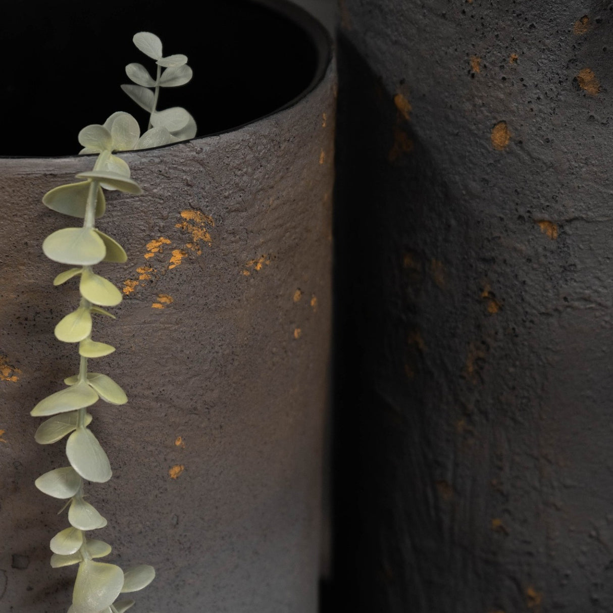Close-up of Libra Interiors Textured Iron Cylinder Planters in mottled blue-grey with gold flecks, one holding a eucalyptus stem against the darker planter.