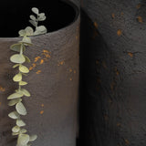 Close-up of Libra Interiors Textured Iron Cylinder Planters in mottled blue-grey with gold flecks, one holding a eucalyptus stem against the darker planter.