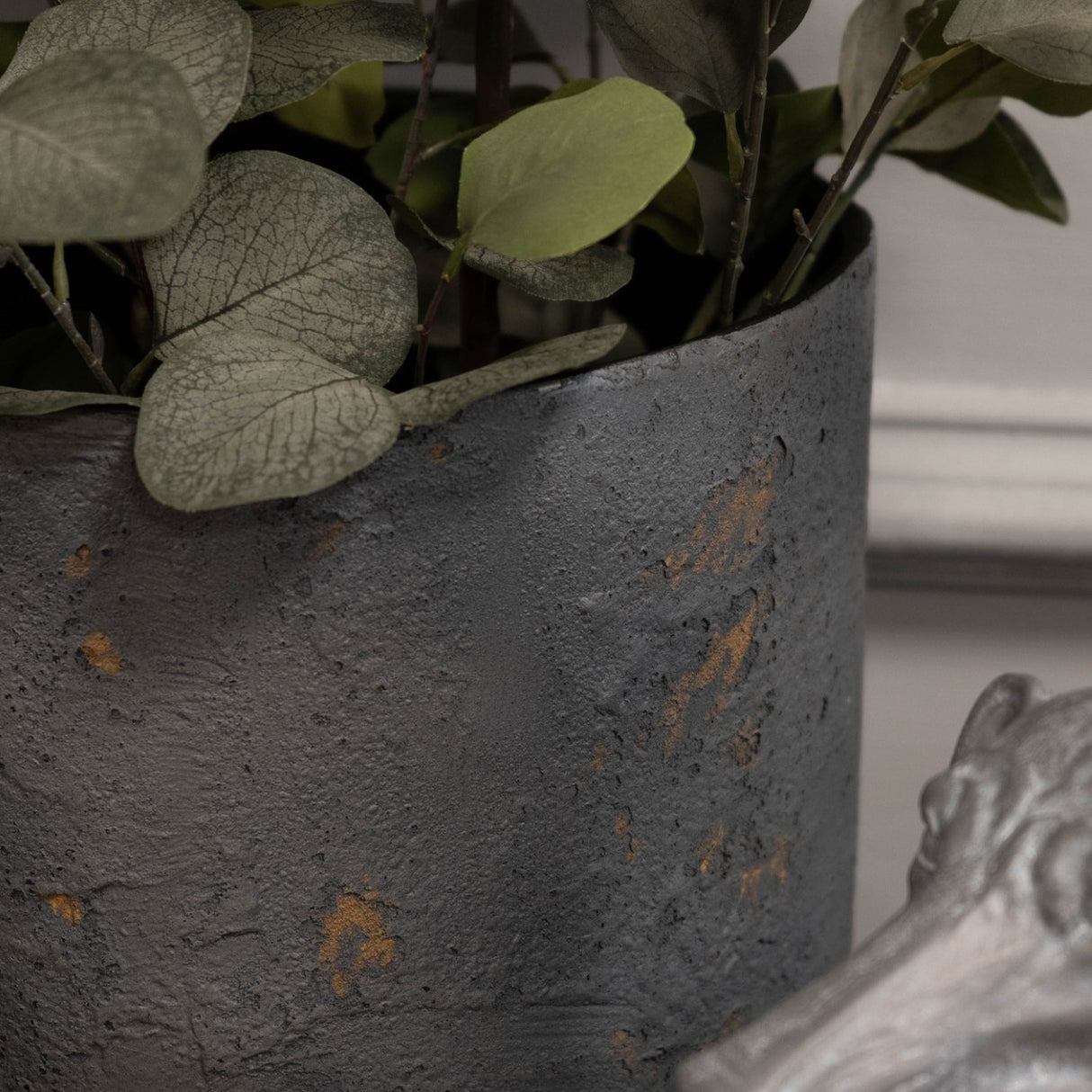 Close-up of Libra Interiors Textured Iron Cylinder Planter in mottled blue-grey with rustic gold flecks, holding eucalyptus stems beside a silver figurine.