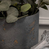 Close-up of Libra Interiors Textured Iron Cylinder Planter in mottled blue-grey with rustic gold flecks, holding eucalyptus stems beside a silver figurine.