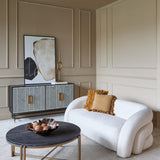Libra Interiors cream velvet wraparound 2-seater sofa with curved design, styled with mustard cushions beside a patterned sideboard and dark round coffee table.