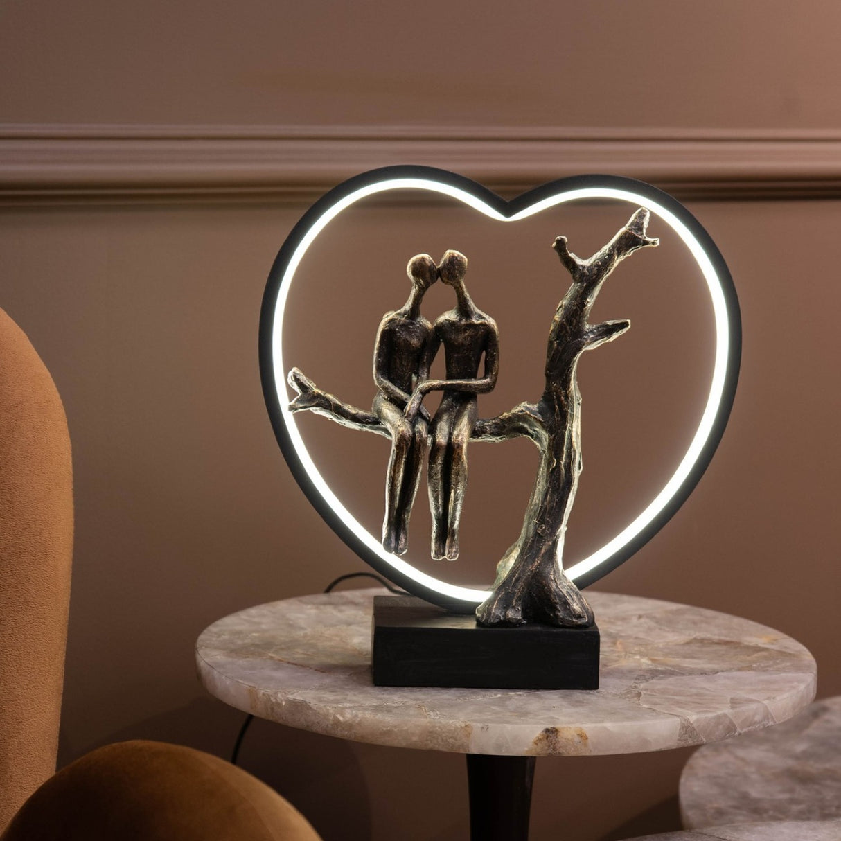 Libra Interiors Antique Bronze Couple LED lighting accessory with resin figures on a branch inside a lit black heart frame, shown on a marble side table.