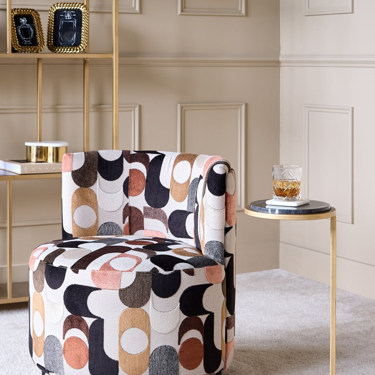 Libra Interiors Patterned Velvet Swivel Armchair in multicoloured retro geometric print, styled beside a gold marble side table and shelving unit.