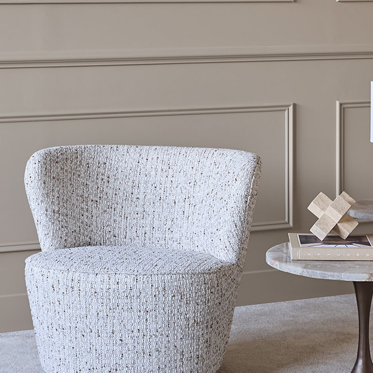 Libra Interiors Textured Swivel Fabric Armchair in natural flecked weave, with rounded low back, set beside a round marble side table against panelled walls.