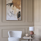 Libra Interiors Textured Swivel Fabric Armchair in natural flecked weave beside a marble side table with wood lamp, below abstract art on panelled wall.