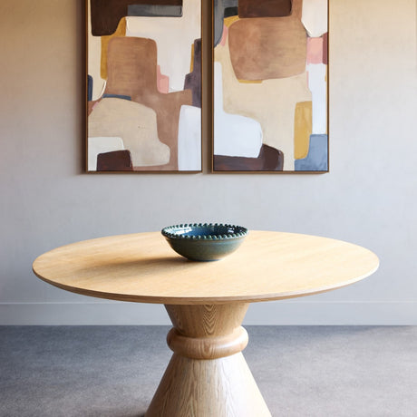 Libra Interiors Round Oak Veneer Pedestal Dining Table in natural finish, with a sculptural twin-cone base and ceramic bowl, set beneath abstract artwork.