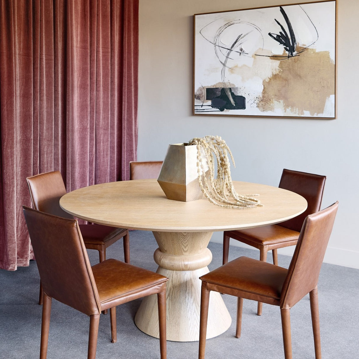 Libra Interiors Round Oak Veneer Pedestal Dining Table in natural finish, styled with brown leather chairs and a brass vase in a dining room setting.