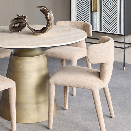 Libra Interiors Curved Back Upholstered Dining Chairs in wheat woven fabric around a round travertine table with brass pedestal in a neutral dining room.