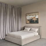 Libra Interiors beige chenille upholstered king bed with padded curved headboard and wraparound base, styled in a neutral bedroom with drapes and abstract art.