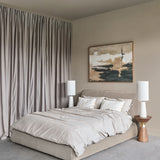 Libra Interiors beige chenille upholstered king bed with padded curved headboard, styled in a neutral bedroom with grey drapes and abstract wall art.