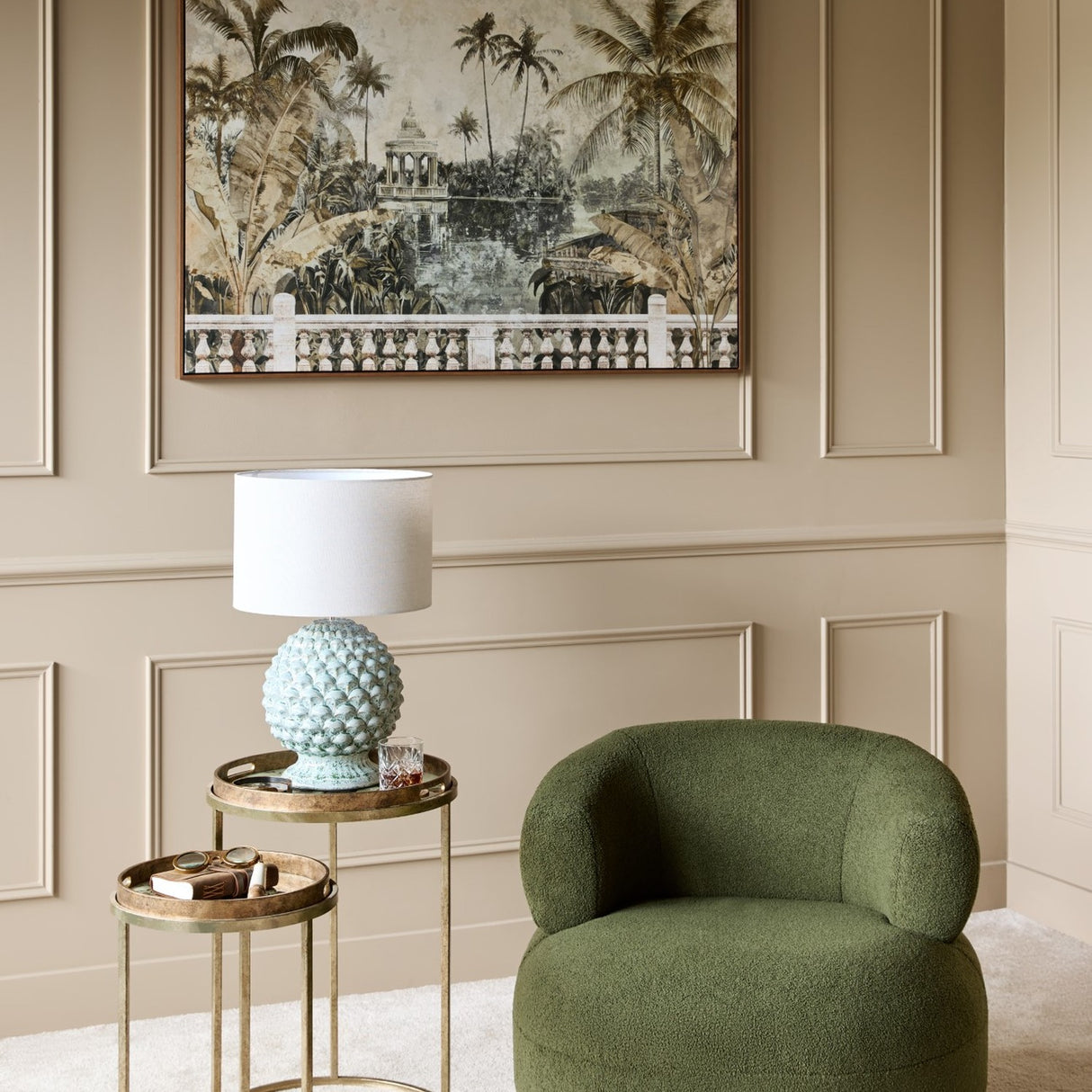 Libra Interiors Glazed Ceramic Artichoke lamp with pale green petal base and white drum shade on a brass side table beside a green bouclé armchair.