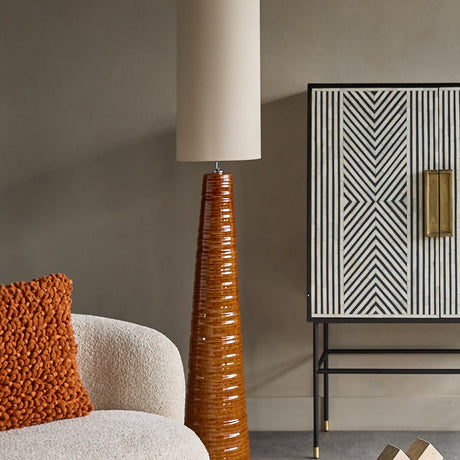 Libra Interiors Ribbed Ceramic Tapered Floor Lamp in terracotta beside a bouclé chair and striped monochrome cabinet in a neutral living room.