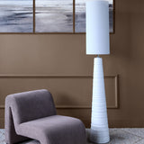 Libra Interiors Ribbed Ceramic Tall Floor Lamp in white with tapered banded base and cylindrical fabric shade, beside a grey corduroy chair in a living room.