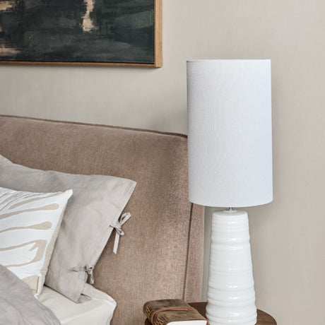 Libra Interiors Ribbed Ceramic Tall Table Lamp in white with a tapered ribbed base and cylindrical fabric shade on a bedside table beside an upholstered bed.