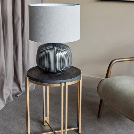 Libra Interiors Ribbed Ceramic Table Lamp in charcoal with grey fabric drum shade, on a round dark wood side table with brass frame beside an armchair.