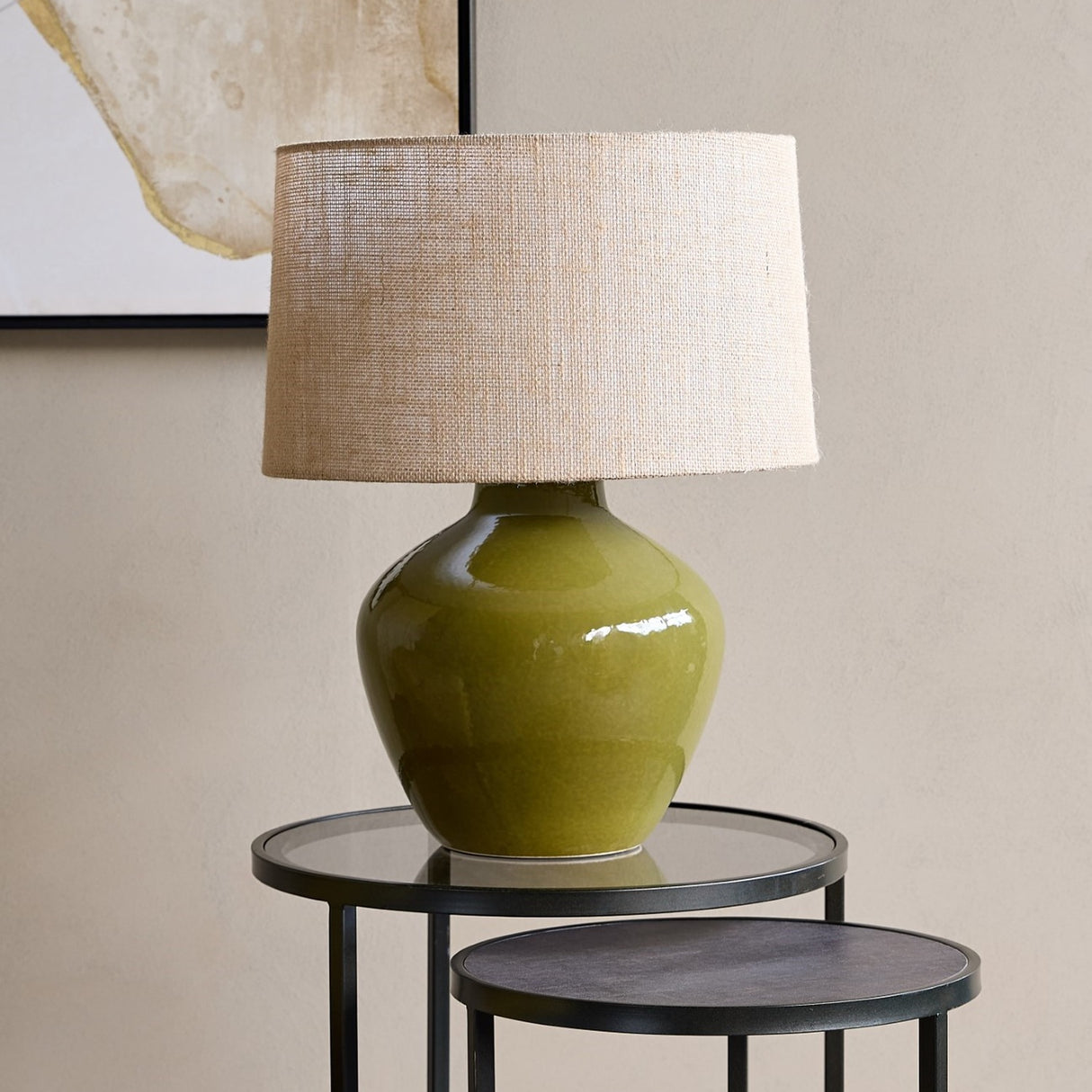 Libra Interiors Glazed Ceramic Hessian Table Lamp in green with a rounded mossy glazed base and natural woven drum shade, on a black side table.
