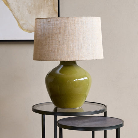 Libra Interiors Glazed Ceramic Hessian Table Lamp in green with a rounded mossy glazed base and natural woven drum shade, on a black side table.