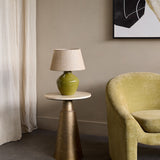 Libra Interiors Glazed Ceramic Hessian Table Lamp in green with urn-shaped base and natural shade, on a brass side table beside a green armchair.