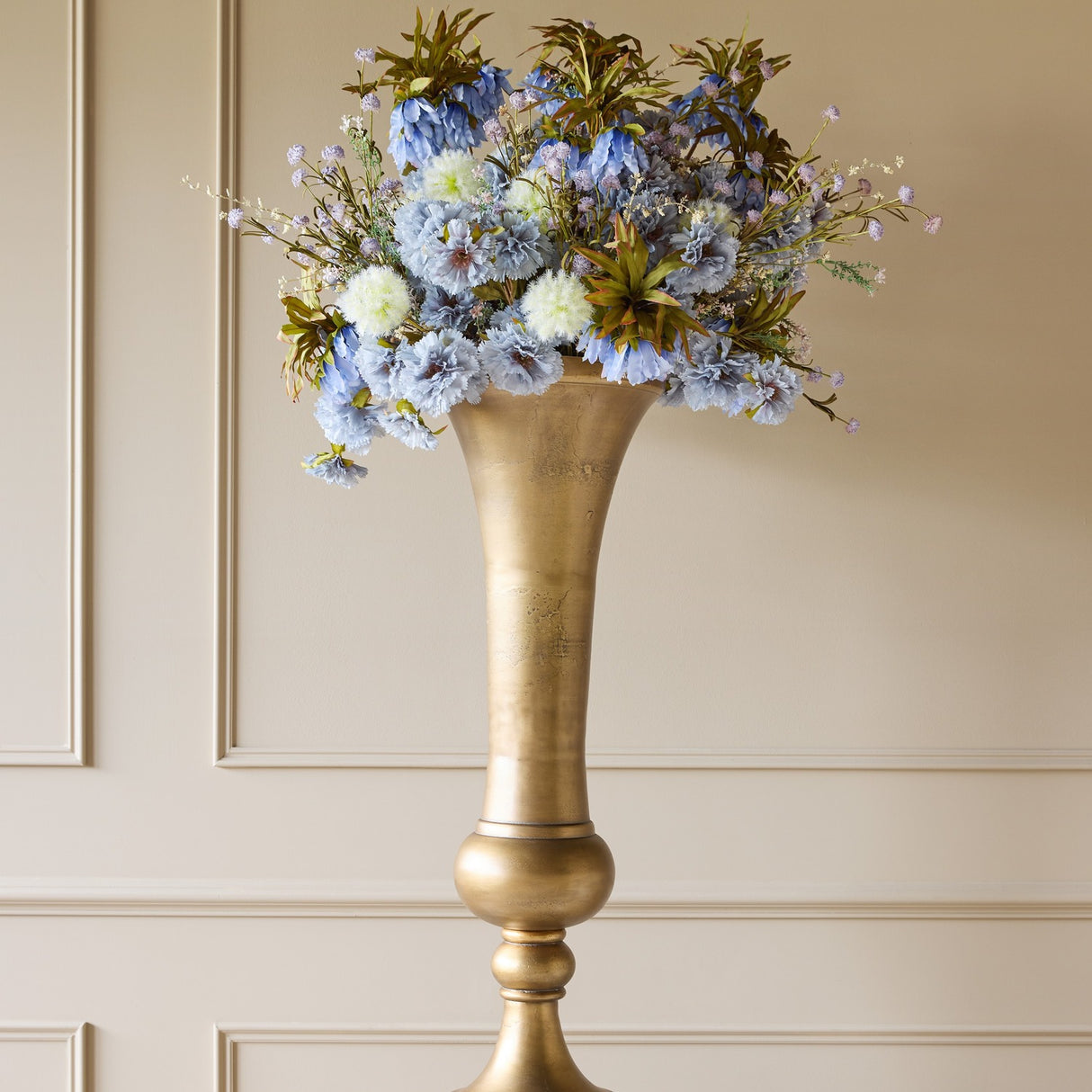 Libra Interiors Trumpet Antique Brass Floor Vase with flared mouth and turned pedestal, displaying blue faux florals against a panelled wall.