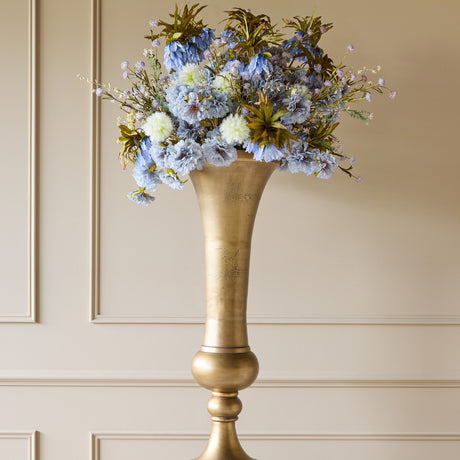 Libra Interiors Trumpet Antique Brass Floor Vase with flared mouth and turned pedestal, displaying blue faux florals against a panelled wall.