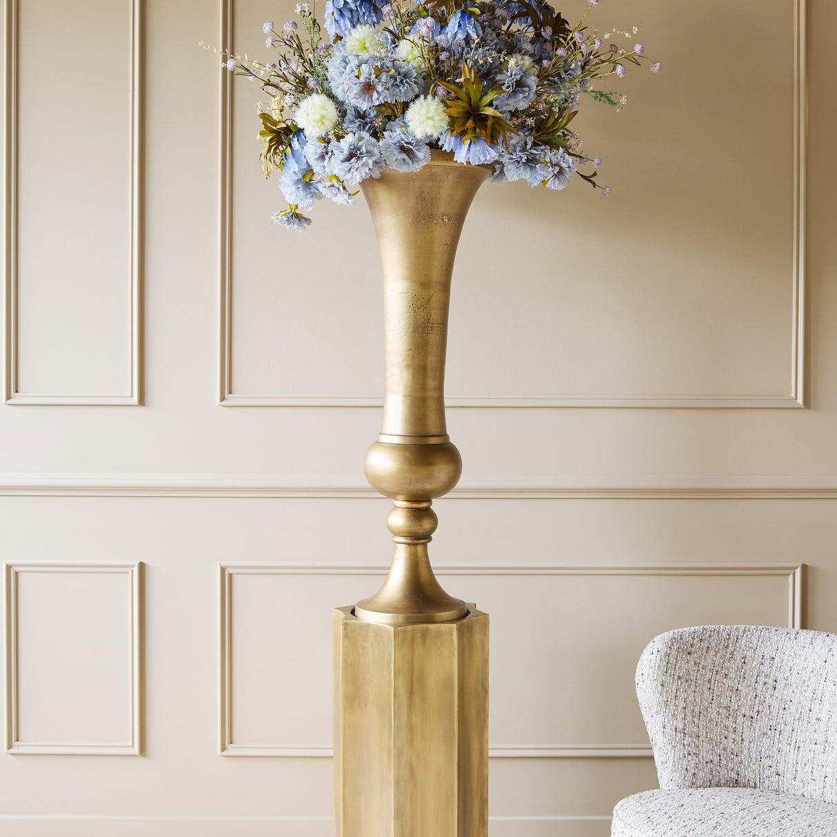 Libra Interiors Trumpet Antique Brass Floor Vase on a hexagonal plinth, filled with blue and cream faux florals, set against a panelled wall.
