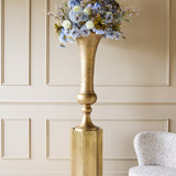 Libra Interiors Trumpet Antique Brass Floor Vase on a hexagonal plinth, filled with blue and cream faux florals, set against a panelled wall.