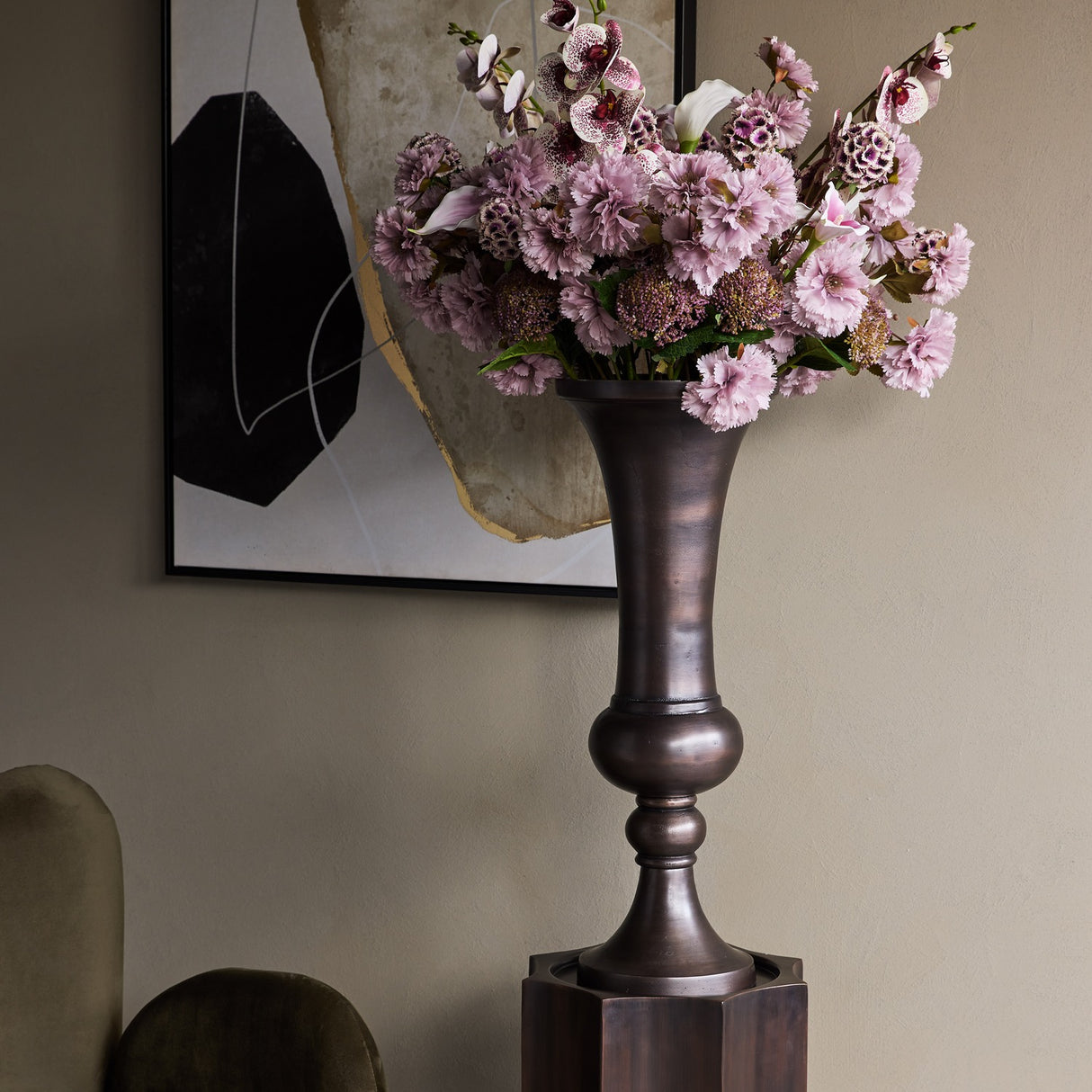 Libra Interiors Trumpet antique bronze aluminium vase with turned baluster stem, holding pink carnations and orchids beside abstract wall art in a hallway.