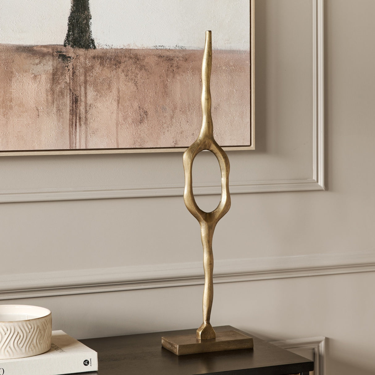 Libra Interiors Cast Aluminium Abstract Sculpture in brass, tall organic form with open oval centre on a square base, styled on a console beneath artwork.