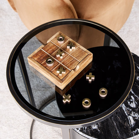 Libra Interiors mango wood noughts and crosses game with brass cross and ring pieces, displayed on a round black glass side table in a living room.