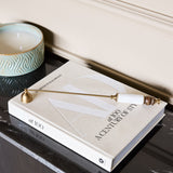 Libra Interiors Brass and Bone Inlay Candle Snuffer resting on an Architectural Digest book beside a patterned ceramic bowl on a dark marble surface.