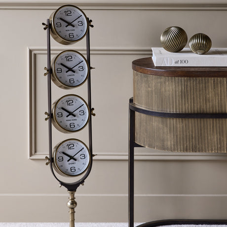 Libra Interiors Brushed Brass Four-Dial World City Floor Clock with vertical New York, London, Hong Kong and Sydney dials beside a fluted console.
