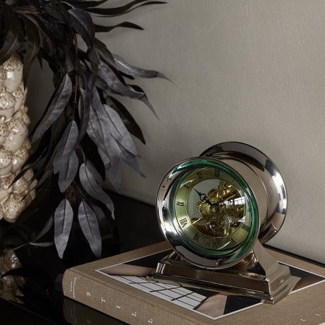 Libra Interiors Skeleton polished nickel mantel clock with domed glass, cream Roman numeral dial and exposed cogs, displayed on a book beside dark foliage.
