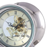 Close-up of the Libra Interiors Skeleton Polished Metal Mantel Clock in nickel, showing exposed gold cogs behind domed glass and a cream Roman numeral dial.