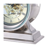 Libra Interiors Skeleton mantel clock close-up showing exposed brass cogs, cream Roman numeral dial and polished nickel case on a stepped aluminium base.