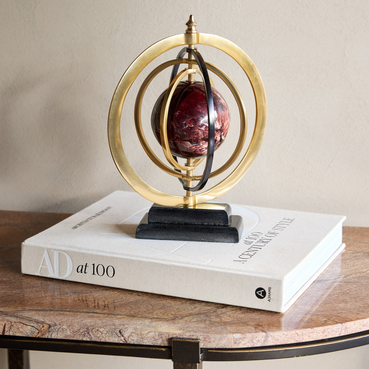 Libra Interiors Marble-Effect Brass Ring Armillary Globe with burgundy resin orb and stepped stone base, styled on an AD at 100 book atop a side table.