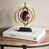 Libra Interiors Marble-Effect Brass Ring Armillary Globe with burgundy resin orb and stepped stone base, styled on an AD at 100 book atop a side table.