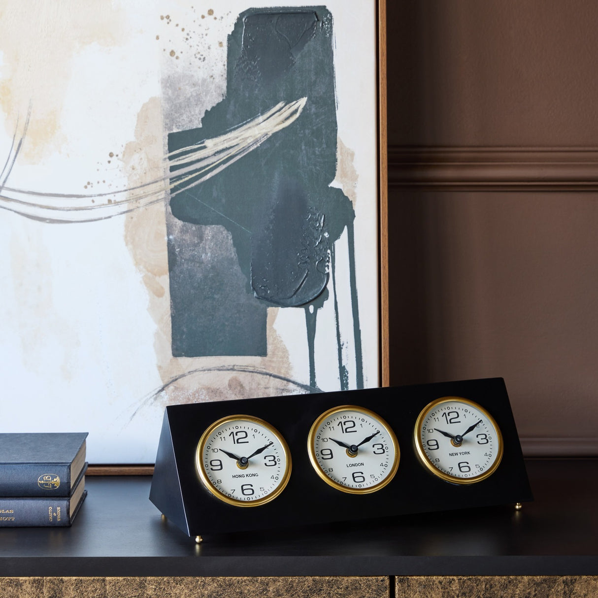 Libra Interiors Matt Black Three Dial Mantel Clock with gold bezels showing Hong Kong, London and New York, on a console beside books and abstract art.