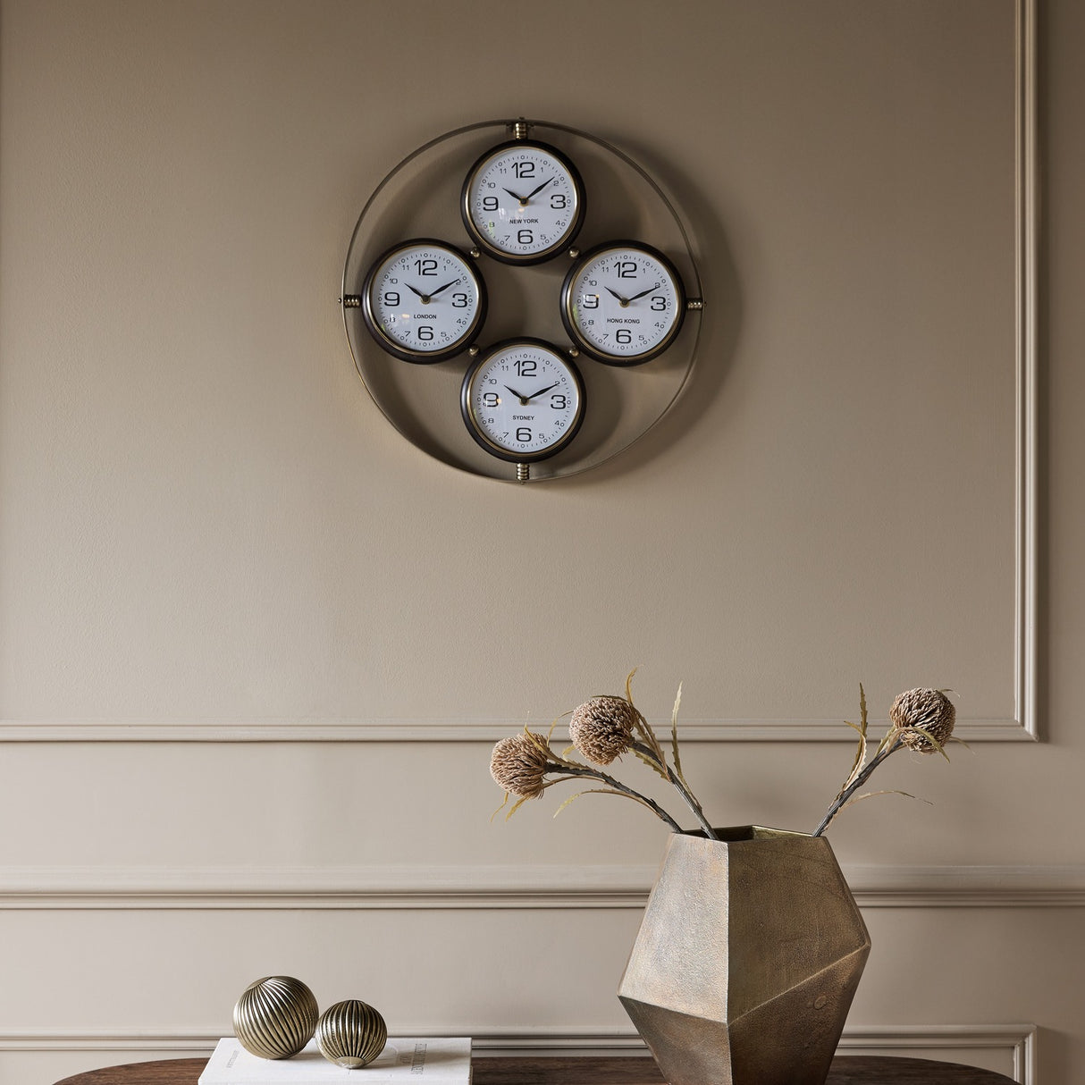Libra Interiors Brushed Brass 4-Dial World City Wall Clock with London, New York, Hong Kong and Sydney faces, mounted on a neutral panelled wall.