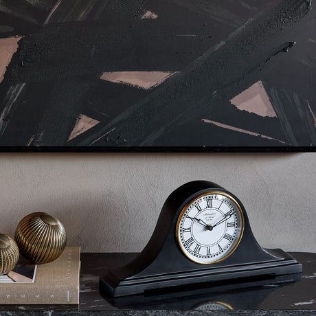 Libra Interiors Matt Black Roman Numeral Mantel Clock on a dark console with brass ornaments and books, beneath a black abstract artwork.