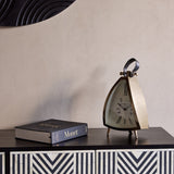 Libra Interiors brushed brass curved front mantel clock with cream Roman numeral dial, black leather handle and splayed feet, styled beside a Monet book.