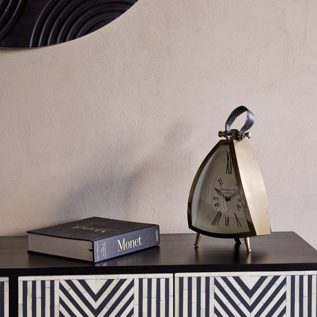 Libra Interiors brushed brass curved front mantel clock with cream Roman numeral dial, black leather handle and splayed feet, styled beside a Monet book.