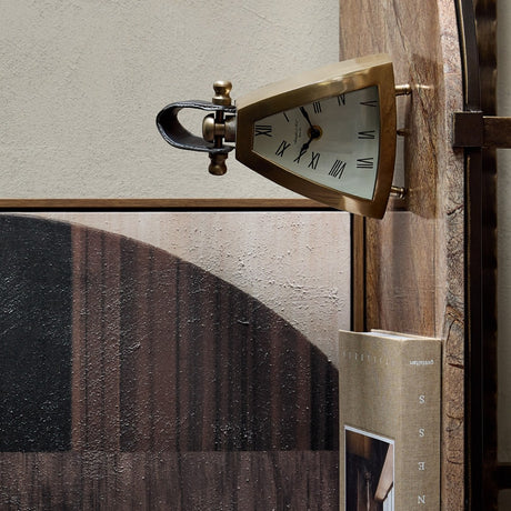 Libra Interiors Brushed Brass Curved Front Mantel Clock with rounded triangular case, white Roman numeral dial and stitched black leather handle, on a shelf.